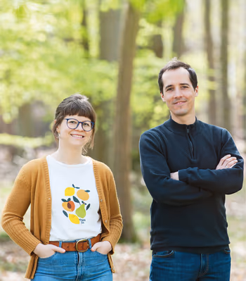 Zwei lächelnde Personen stehen im sonnigen Wald und schauen in die Kamera. Freundliche, authentische Porträtaufnahme in natürlicher Umgebung mit frischem Frühlingslicht.